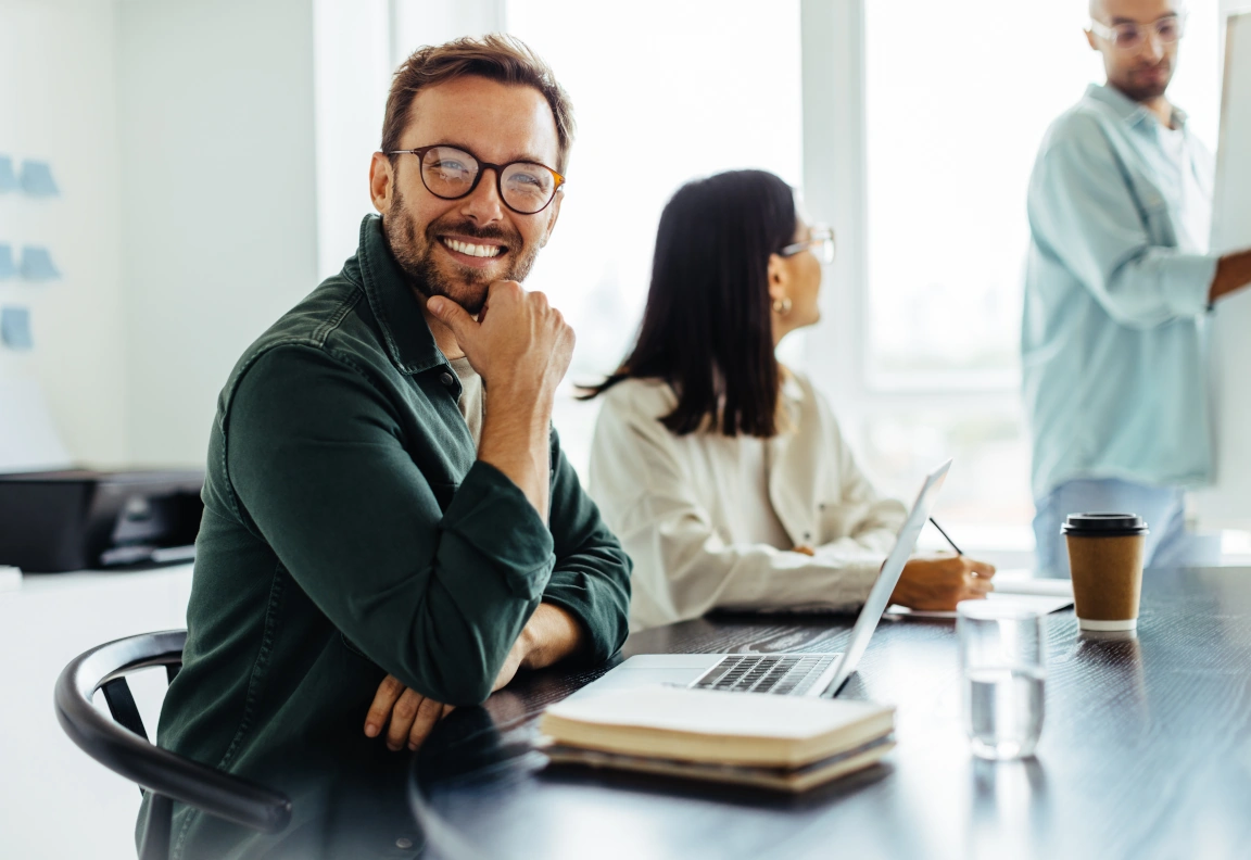 Lachende man achter bureau met laptop vergezeld door twee collega's.