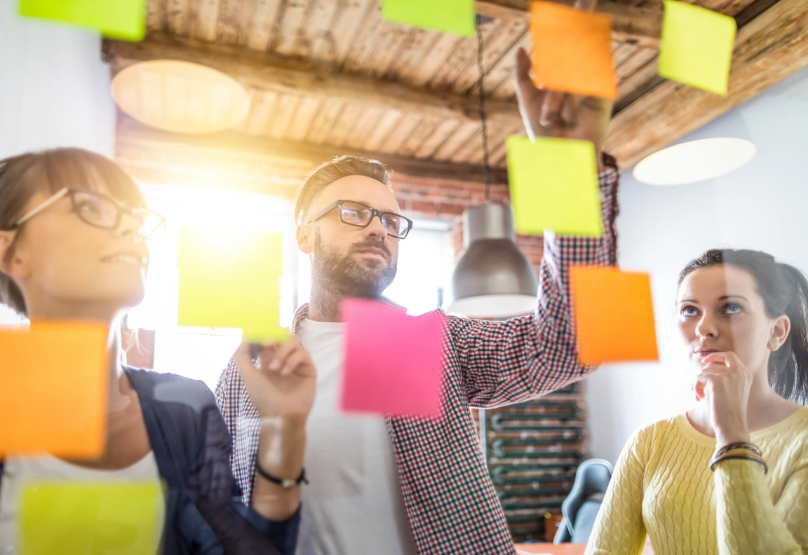 Drie mensen tijdens een brainstormsessie plakken verschillende kleuren post-its op het raam.