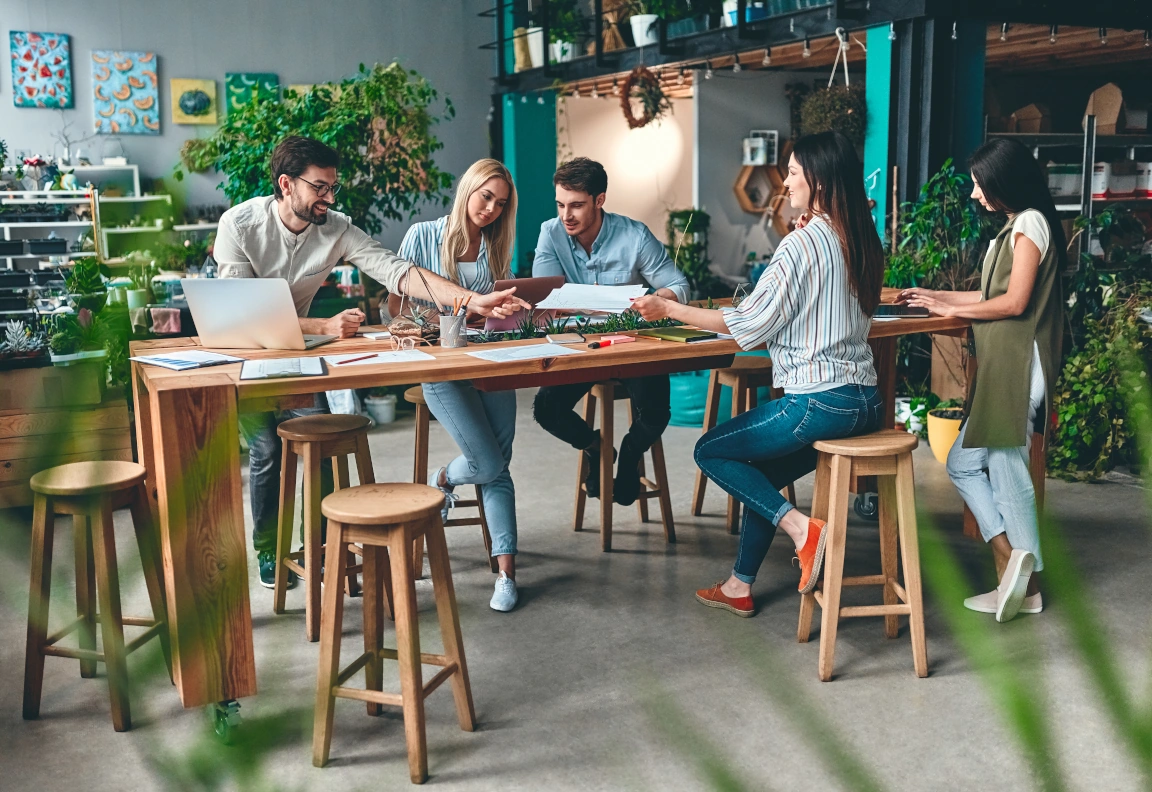 Team van vijf jonge mensen aan een hoge tafel met papieren en laptops samen aan het werk.