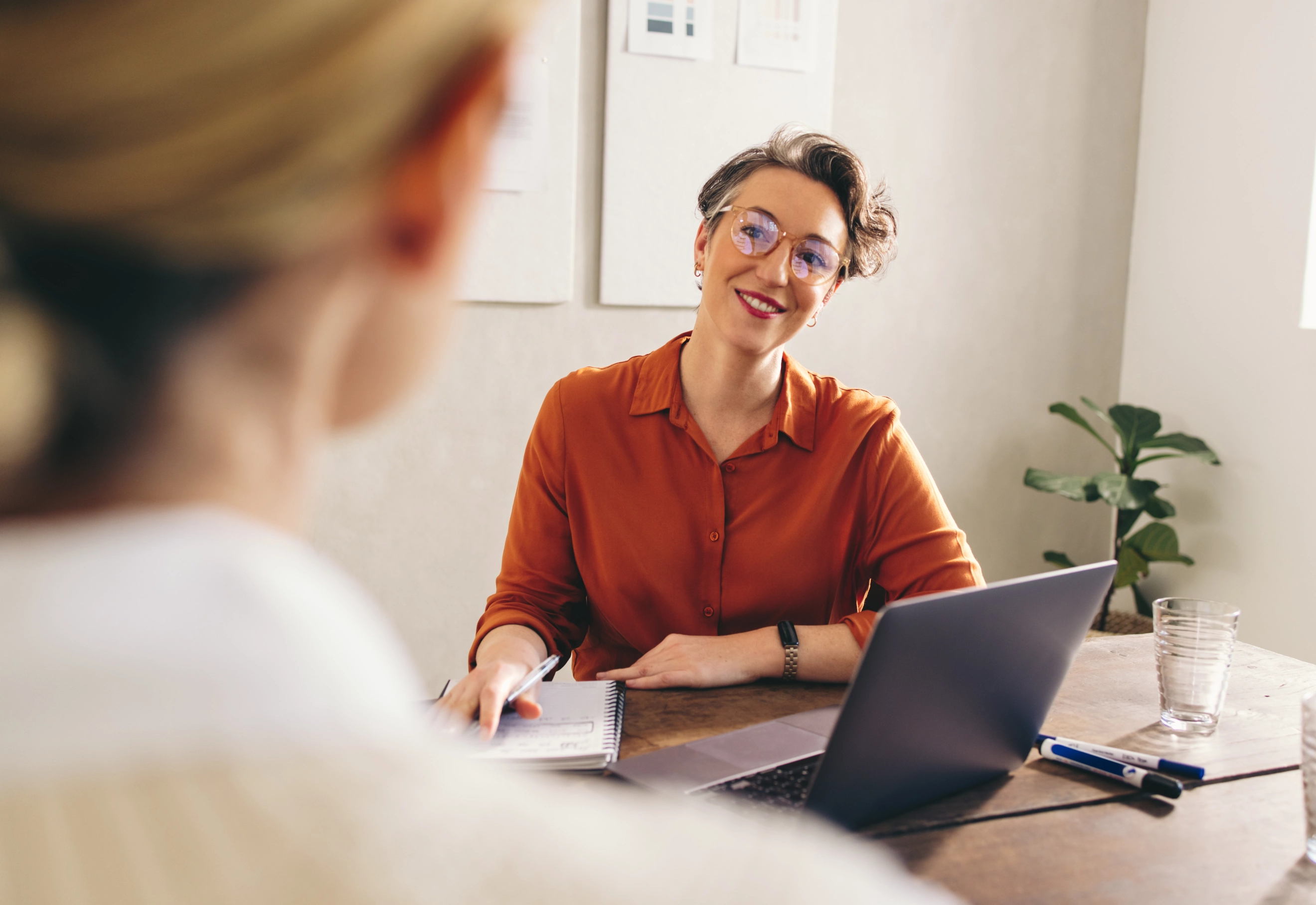 Twee vrouwen in gesprek aan bureau met laptop en papier.